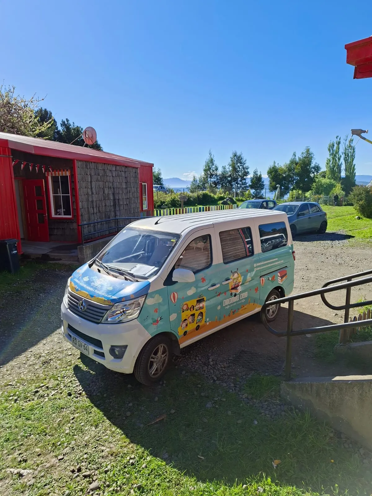 El furgón del Bibliobús Moviendo Sueños estacionado frente a una casa roja de madera, con el lago y los cerros al fondo.