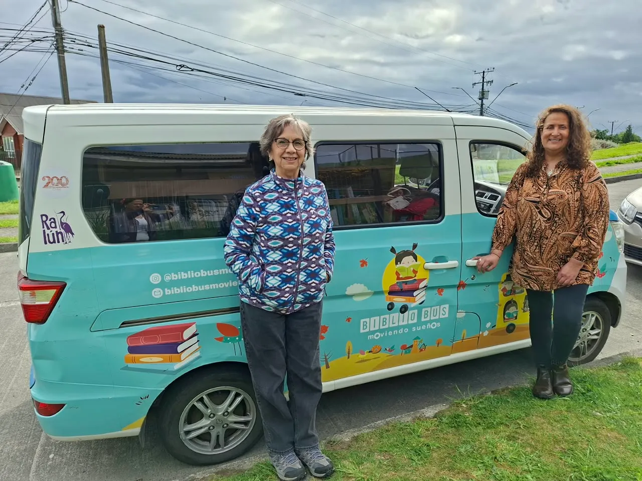 Dos mujeres de pie junto al furgón celeste del Bibliobús Moviendo Sueños, con dibujos de niños y libros en la carrocería.