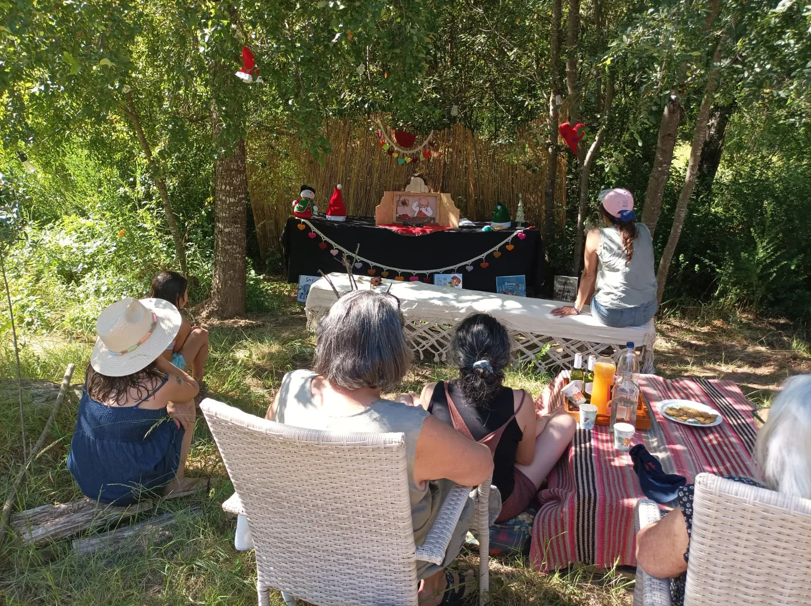 Público sentado en un jardín mira una función de kamishibai sobre una mesa decorada con corazones, bajo los árboles.