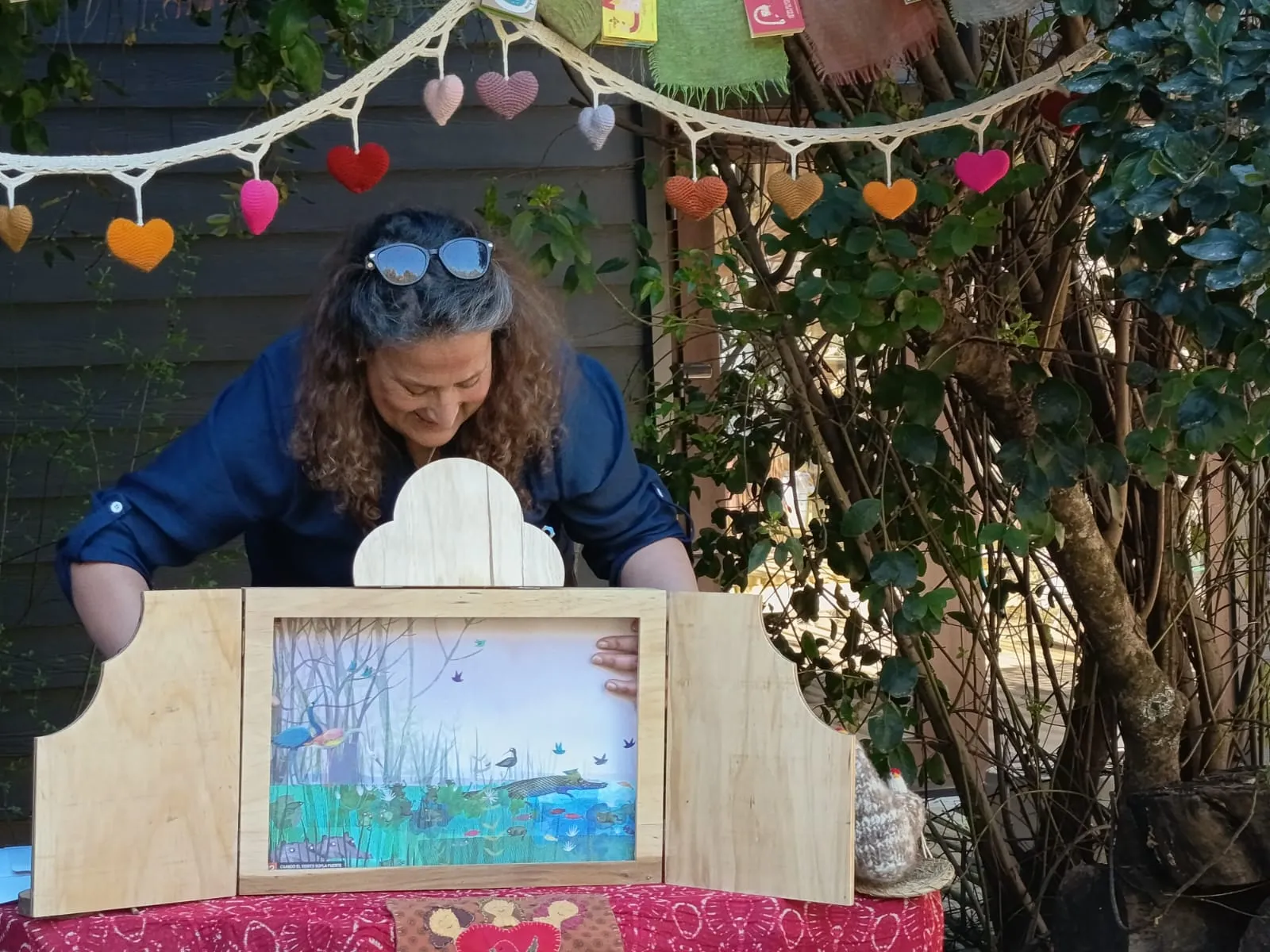 Primer plano de una cuentacuentos sonriendo detrás de su kamishibai de madera, con una lámina ilustrada de humedal y aves, bajo una guirnalda de corazones tejidos.