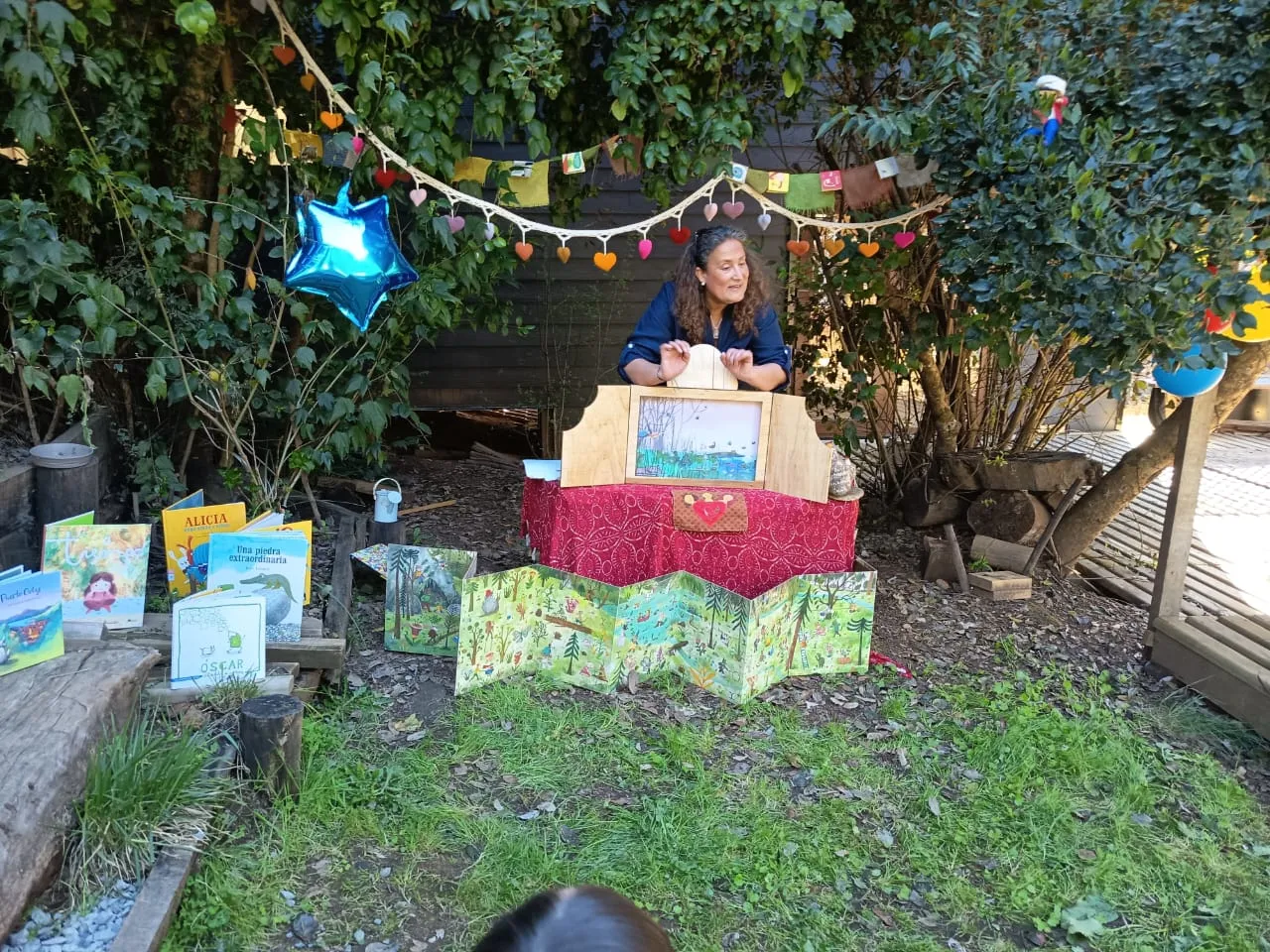 Una cuentacuentos detrás de su kamishibai de madera, con libros infantiles y un biombo ilustrado de bosque sobre el pasto, bajo guirnaldas de corazones de colores.